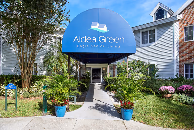 Aldea Green Eagle Senior Living entrance with blue curved sign and tropical plants.