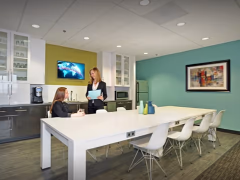 Two professionals converse at a long white table in a modern staff lounge.