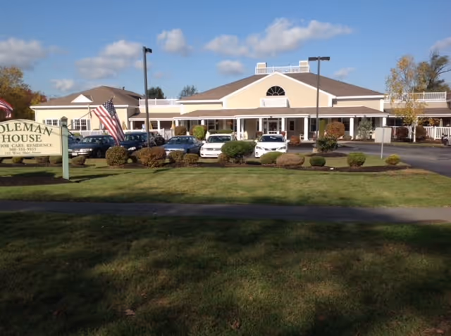 Front exterior view of Coleman House senior care residence building with a parking lot in front, several cars parked, an American flag, and a green lawn with bushes.