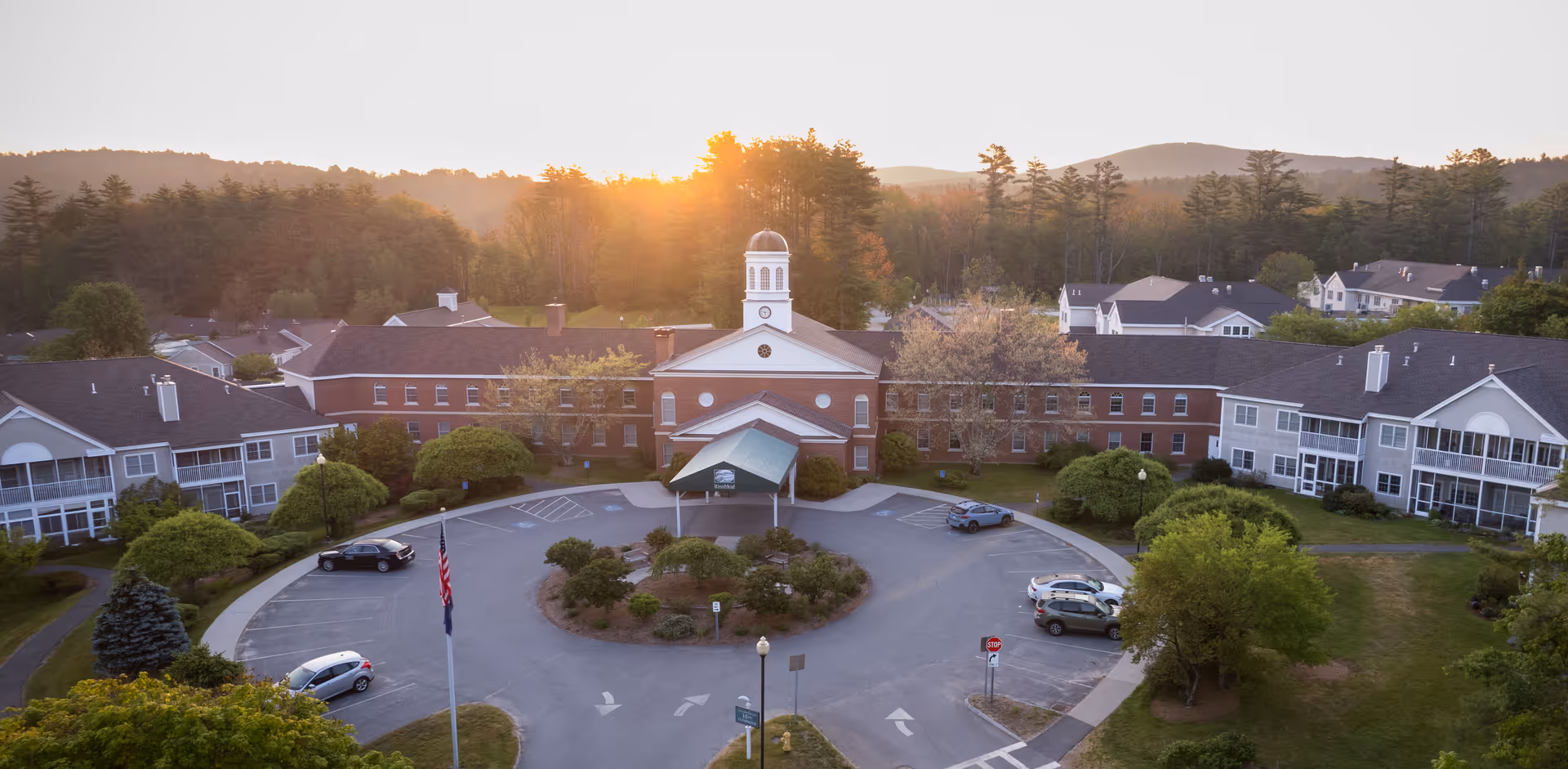 Aerial view of RiverMead's main brick building with a domed cupola, circular driveway, and surrounding residential wings at sunset.