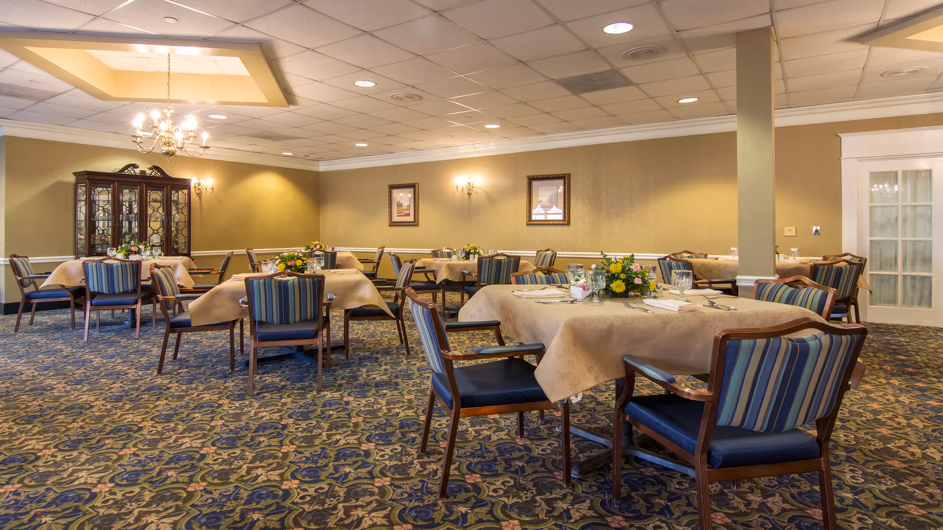 Formal dining room with beige tablecloths, striped chairs, and floral centerpieces.