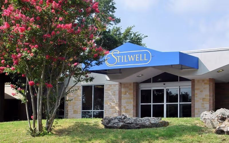 Front entrance of the Stilwell retirement residence featuring a blue awning with "STILWELL", glass doors, stone facade, and a pink-flowering tree on the lawn.