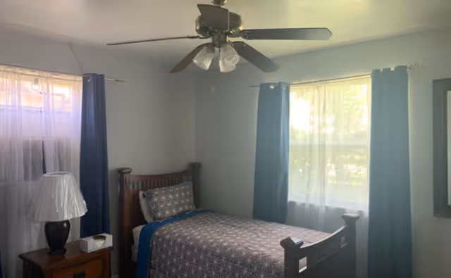 Small furnished bedroom with a single wooden bed, bedside table and lamp, two curtained windows and a ceiling fan.