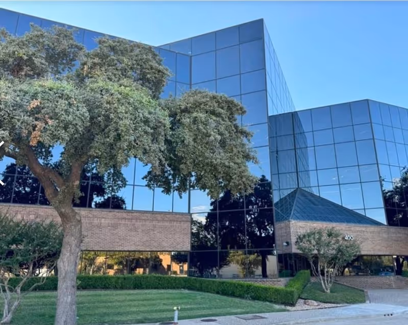 Modern glass-front building with trees and a manicured lawn in front.