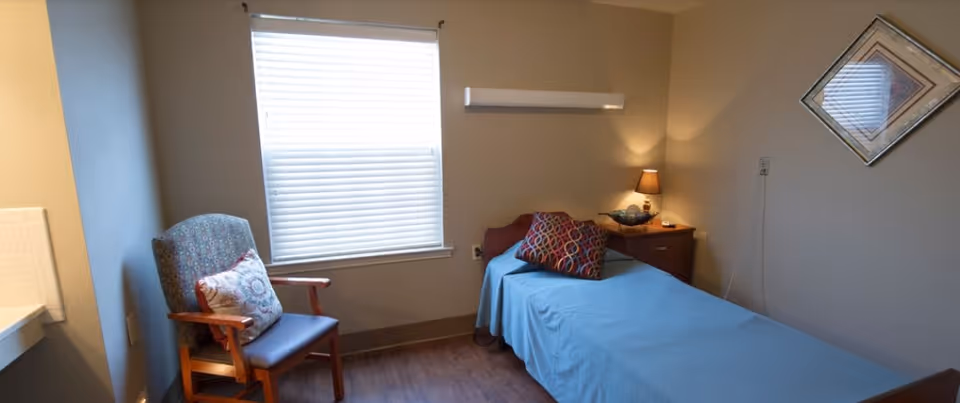 A small bedroom with a single bed covered in a blue sheet and two decorative pillows. Next to the bed is a wooden nightstand with a lamp and a decorative item. A patterned armchair with a cushion is placed near a window with closed blinds. The walls are painted beige, and there is a framed picture hanging on the wall.