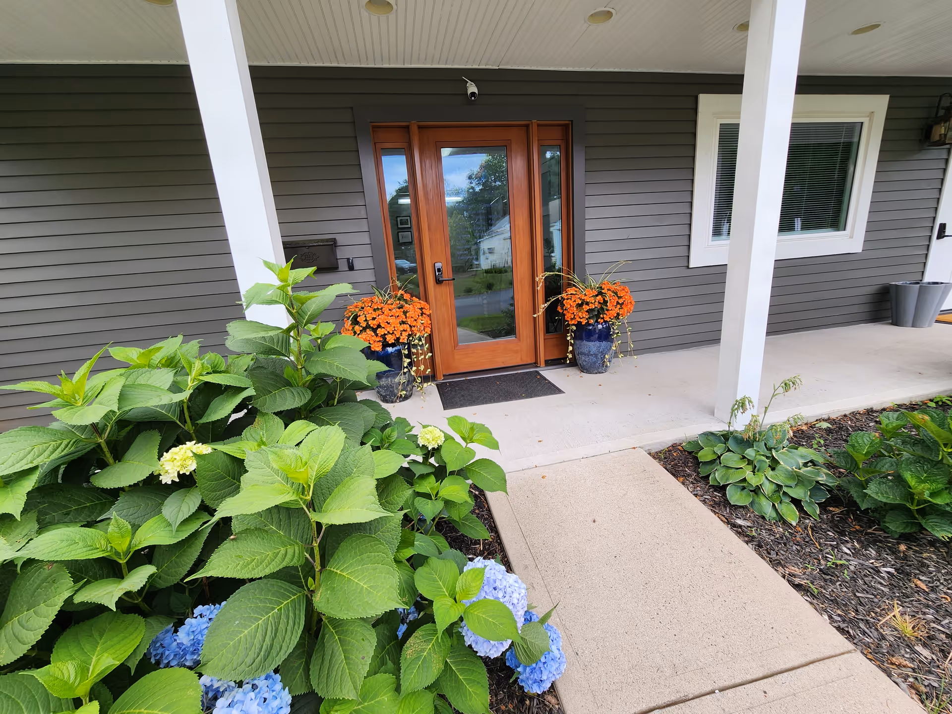 Gray exterior with wooden front door, orange potted flowers, and lush greenery.