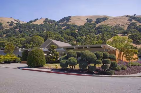A single-story senior living facility with manicured shrubs and rolling hills in the background.
