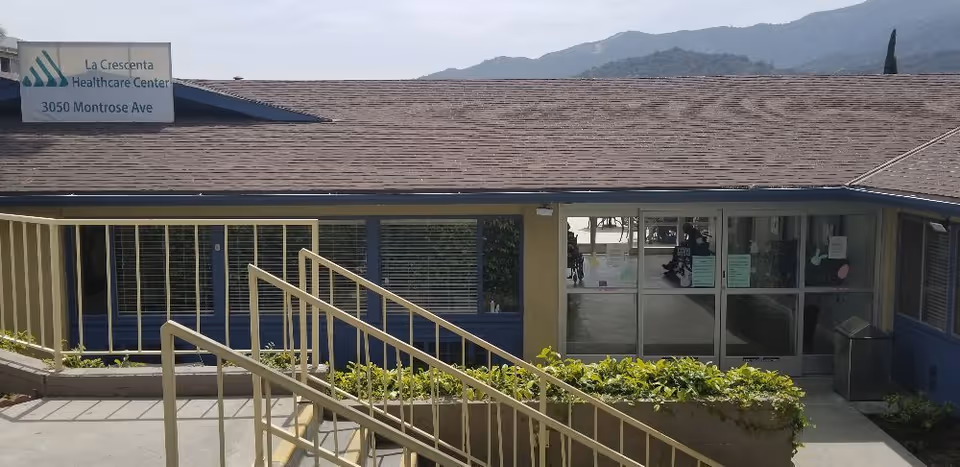 Front entrance of La Crescenta Healthcare Center showing a ramp with handrails, planter, and glass double doors under the building roof.