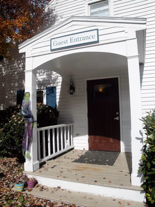 Front porch entrance of a white building with a dark red door and a sign above reading 'Guest Entrance'. There is a small covered porch with white railing, a black outdoor light fixture, and some fallen leaves on the wooden floor. A Halloween decoration of a green-faced figure is hanging on the left porch post, and there are some potted plants on the ground near the steps.