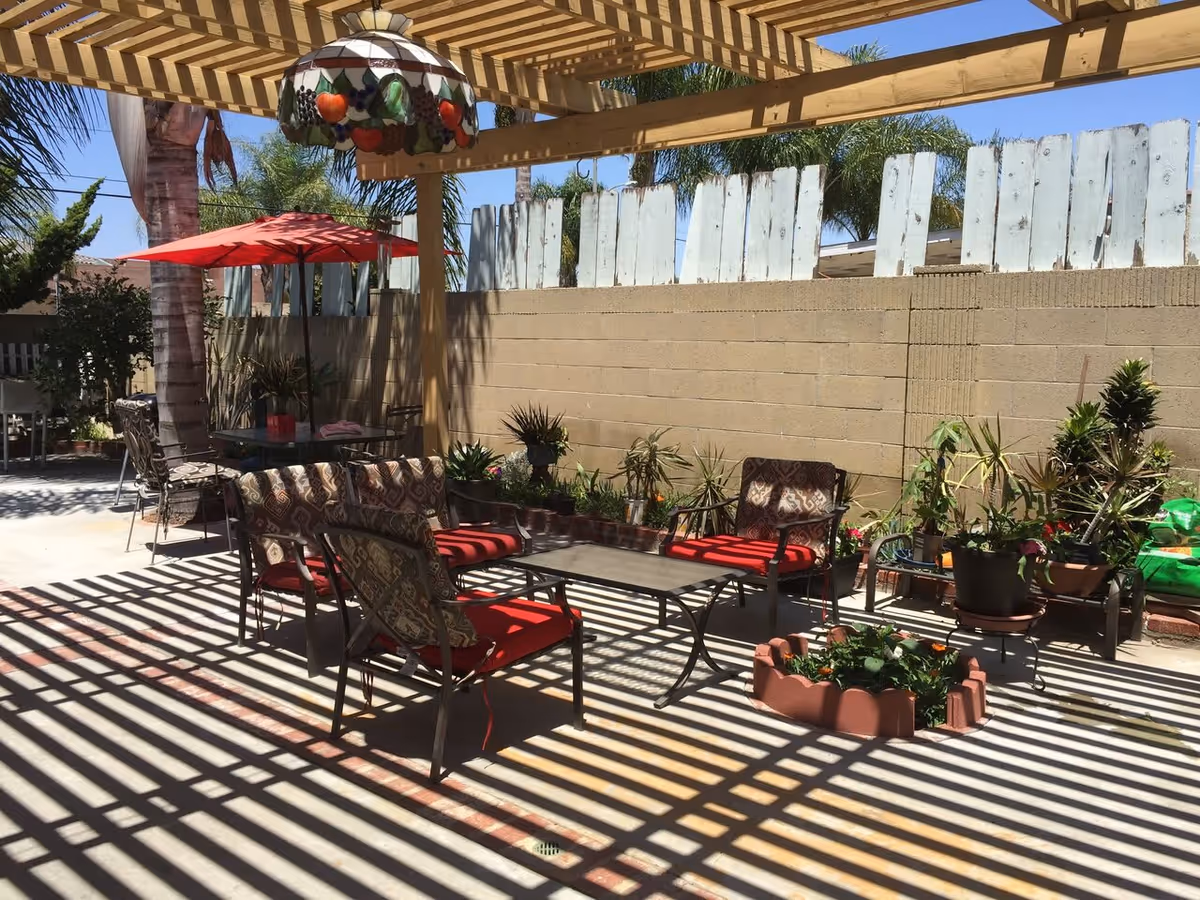 Shaded backyard patio with a pergola casting striped shadows over seating with red cushions, potted plants, and a red umbrella.
