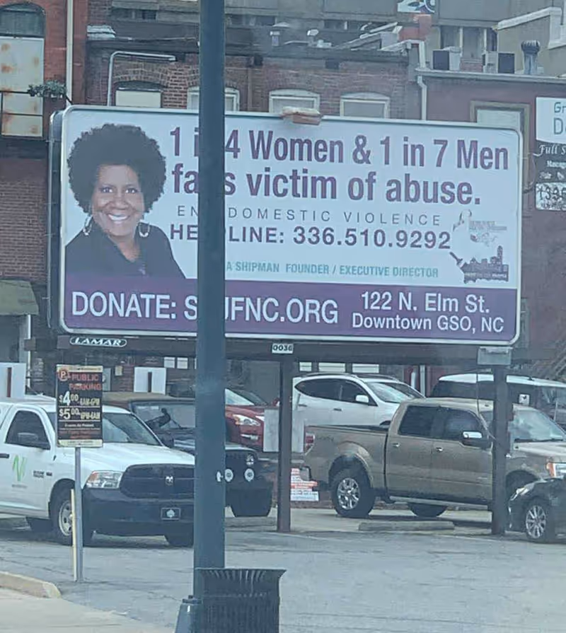 A large street billboard displays a portrait and text about abuse victims, with parked cars nearby.