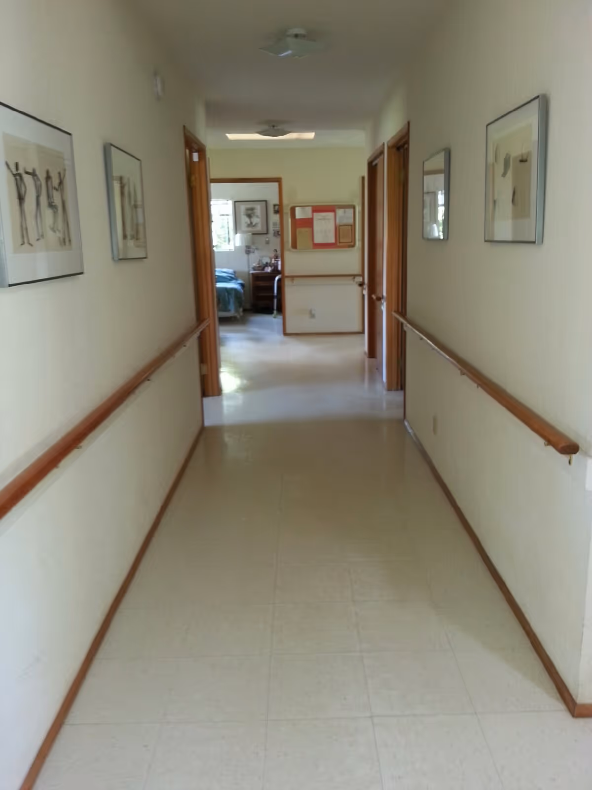 A clean, well-lit hallway in a care home with handrails on both sides and framed artwork hanging on the walls. At the end of the hallway, an open door reveals a bedroom with a bed, nightstand, and window.