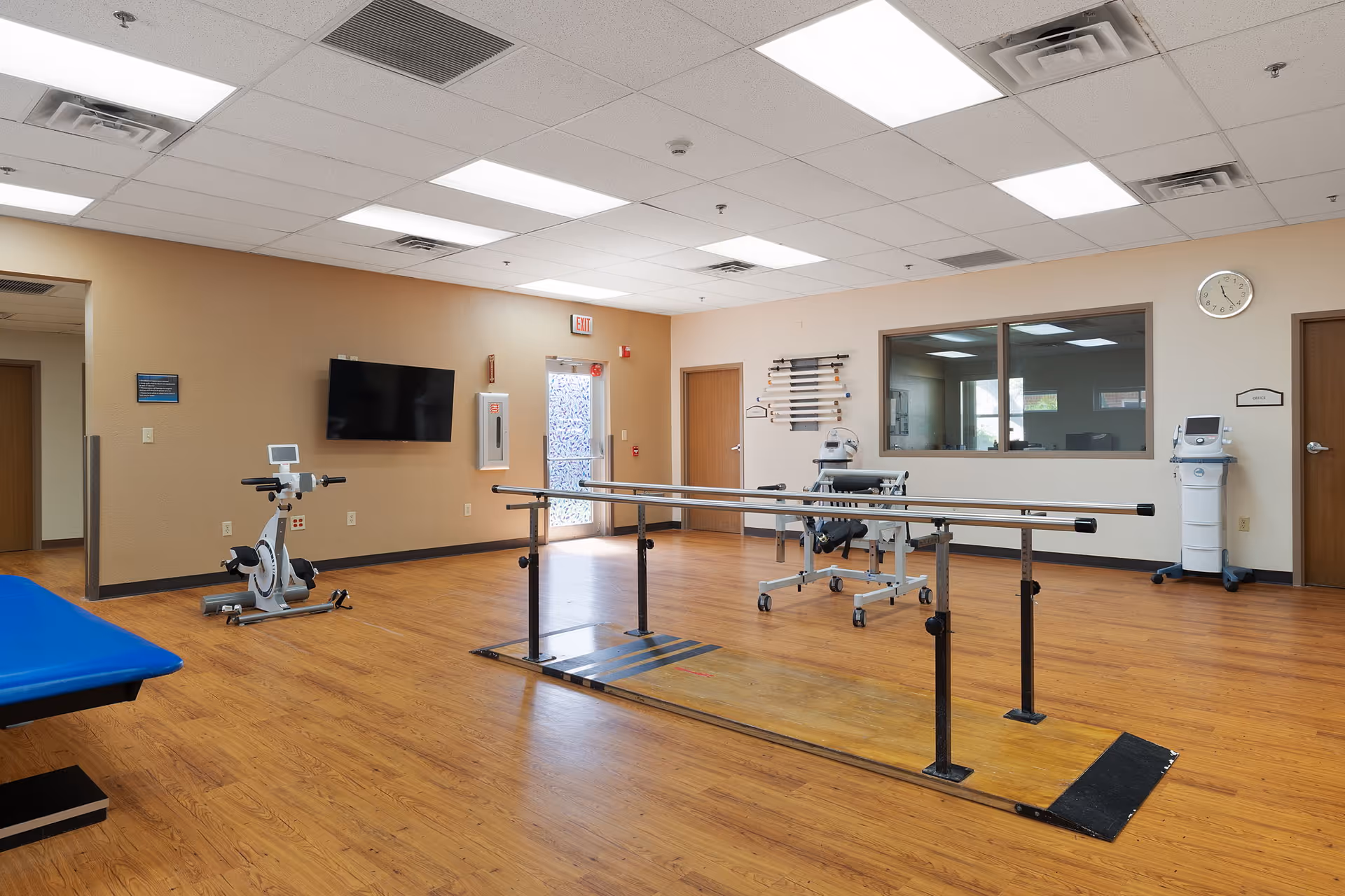 A spacious rehabilitation room with wooden flooring and beige walls. The room contains parallel bars for walking exercises, a stationary exercise bike, a blue padded therapy table, and various medical equipment. There is a wall-mounted TV, an exit door with a frosted glass panel, and a large window looking into another room. The ceiling has fluorescent lighting panels.