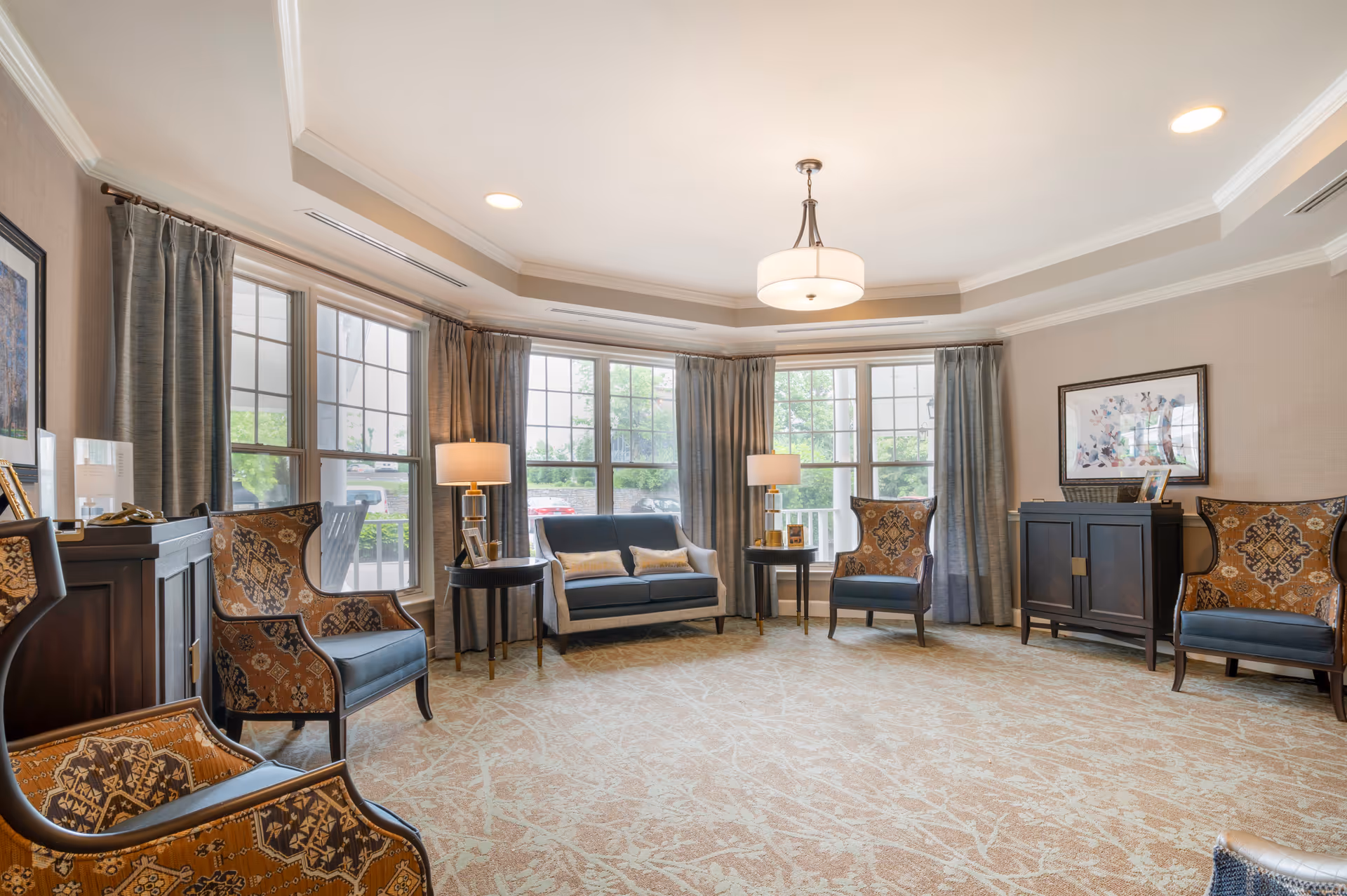 Bright lounge area with sofas, chairs, lamps, and large windows in a senior living facility.