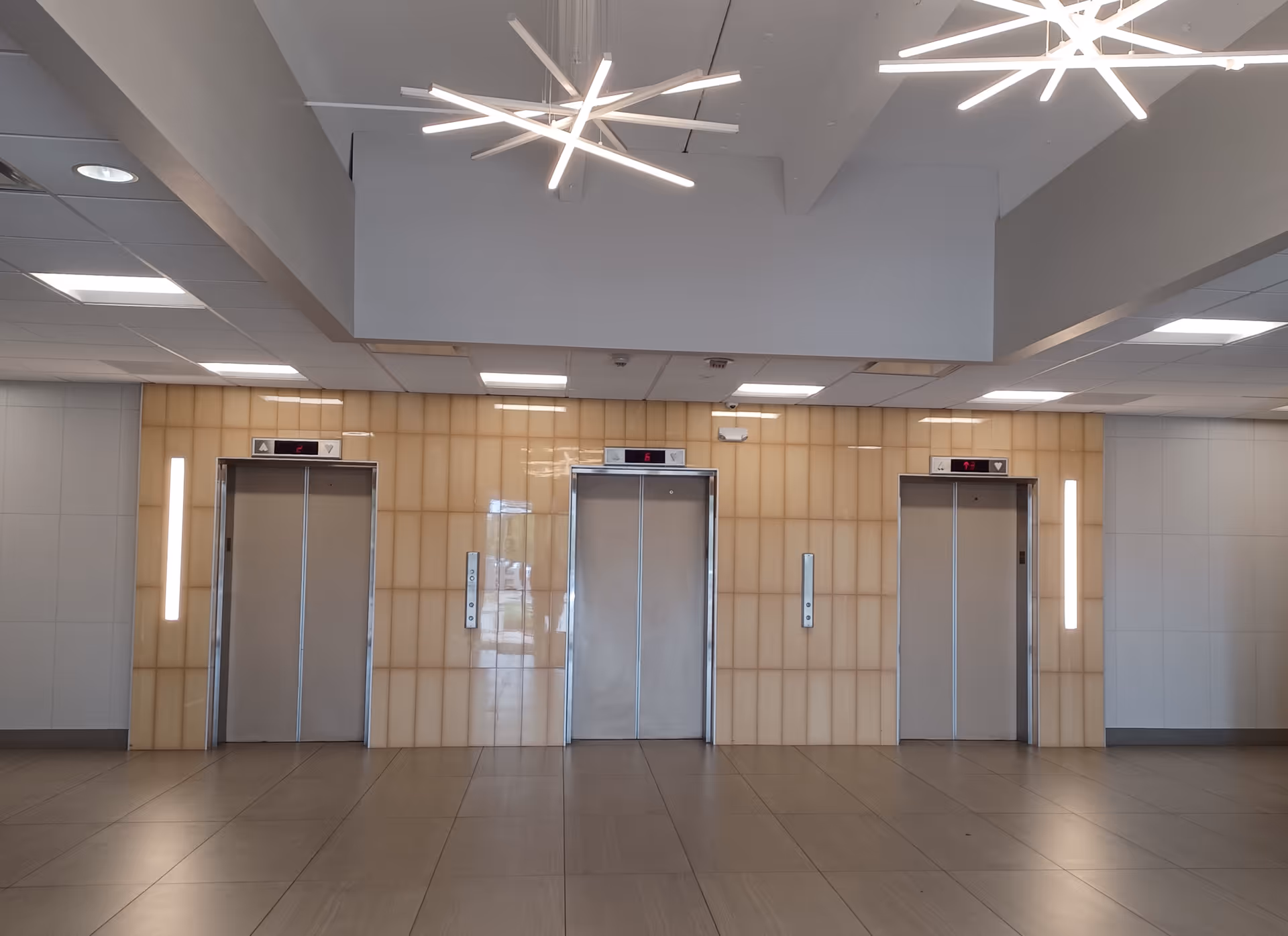Interior of a modern elevator lobby with three doors, beige tiled wall, and bright overhead lighting.