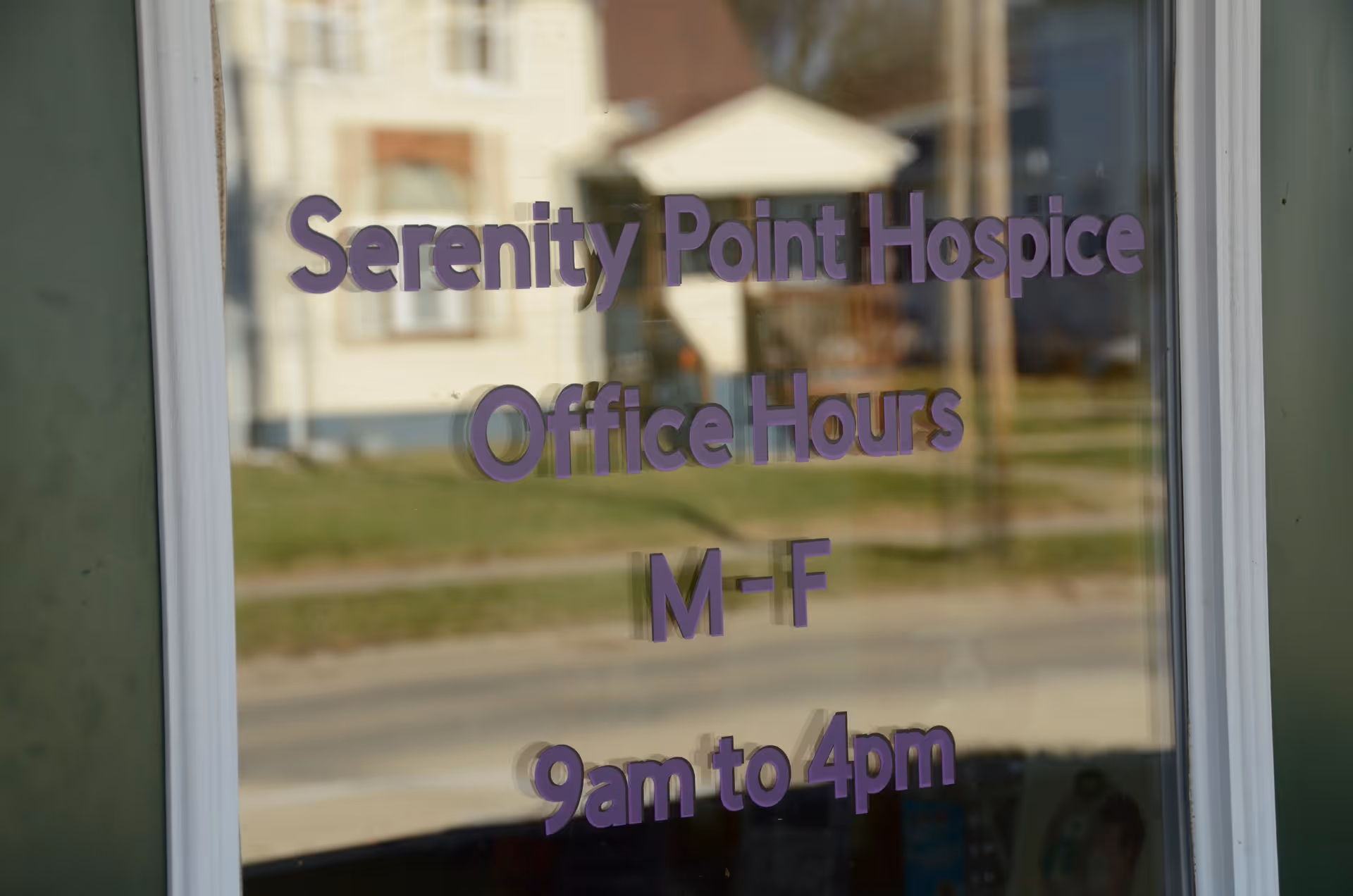 Glass window sign displays Serenity Point Hospice office hours, M-F, visible reflections of surrounding buildings.