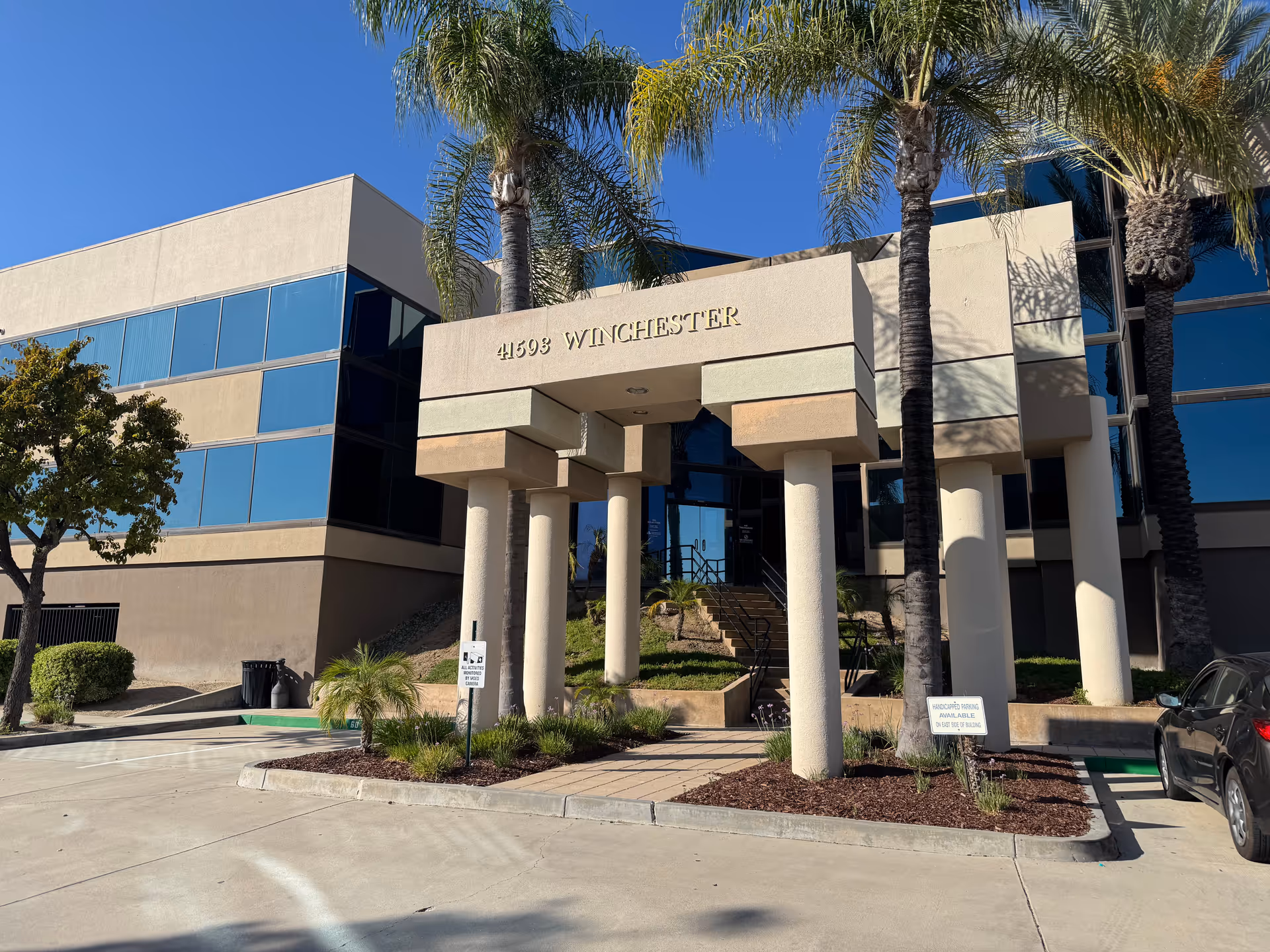 Exterior view of a modern building entrance with beige columns and palm trees.