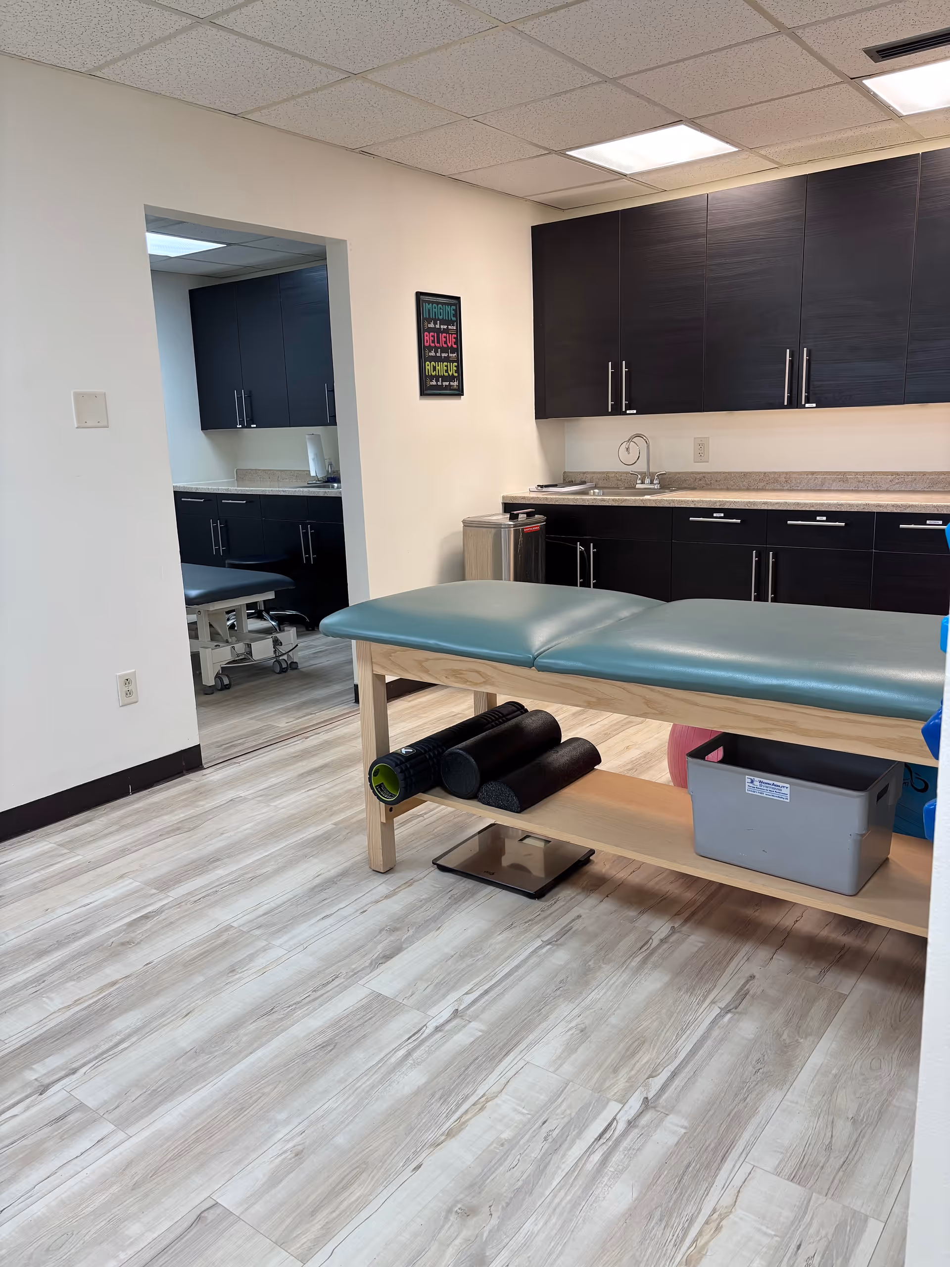 A therapy room with a padded examination table, dark cabinets, and rolled exercise mats.
