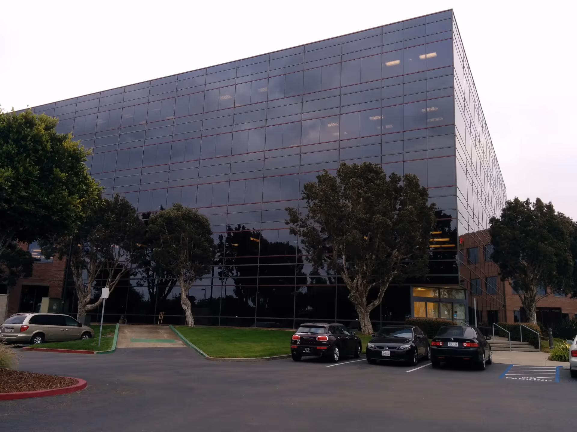 Glass-front Maxim Healthcare building with trees, a parking lot, and parked cars.