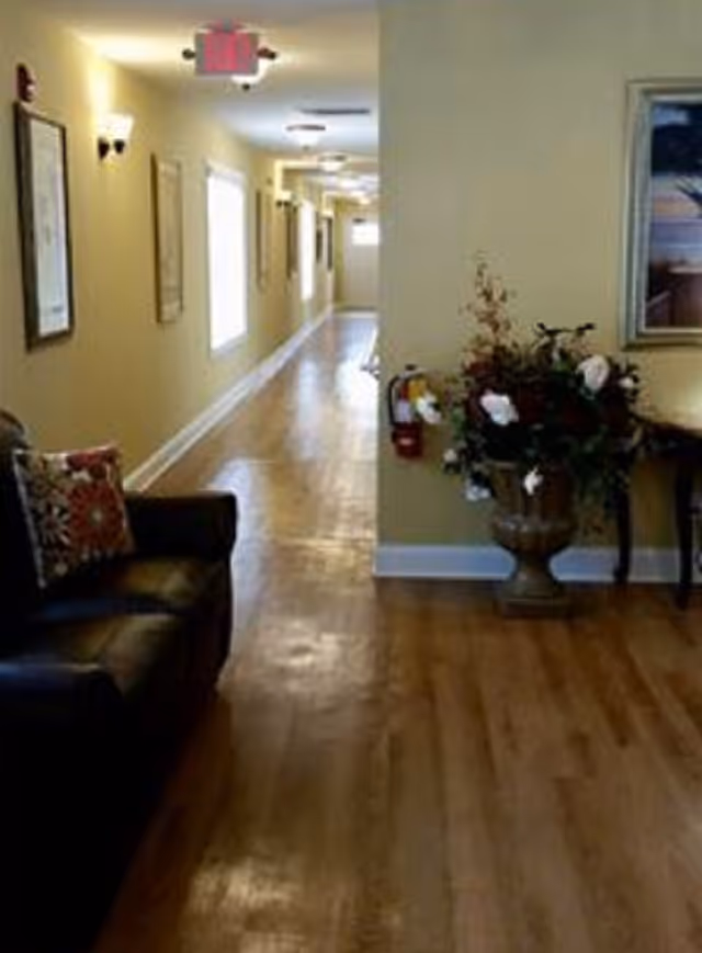 Long indoor hallway in a senior living facility with seating, wall art, and a floral arrangement.