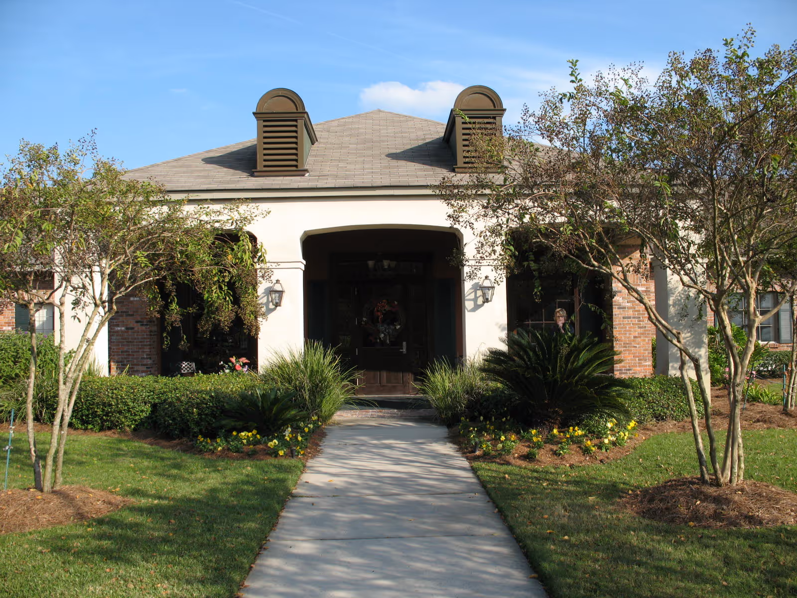 Front exterior view of a single-story building with a pathway leading to the entrance. The building has a light-colored facade with brick accents, two dormer vents on the roof, and is surrounded by neatly trimmed bushes, small trees, and flower beds under a clear blue sky.