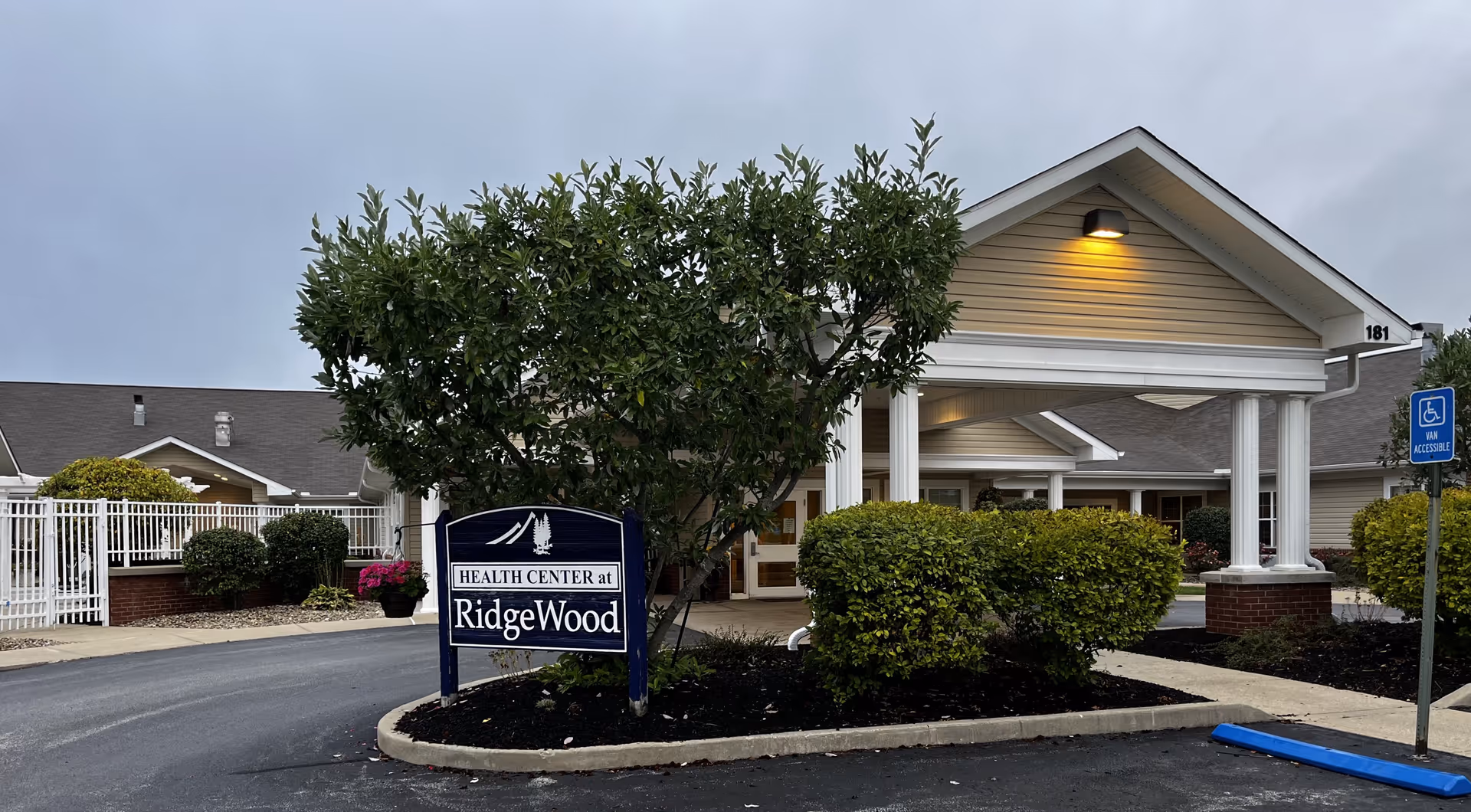 Exterior view of Ridgewood Health Campus building entrance with a covered drop-off area, surrounded by bushes and a tree. A sign in front reads 'Health Center at RidgeWood'. There is a handicapped parking sign visible on the right side.
