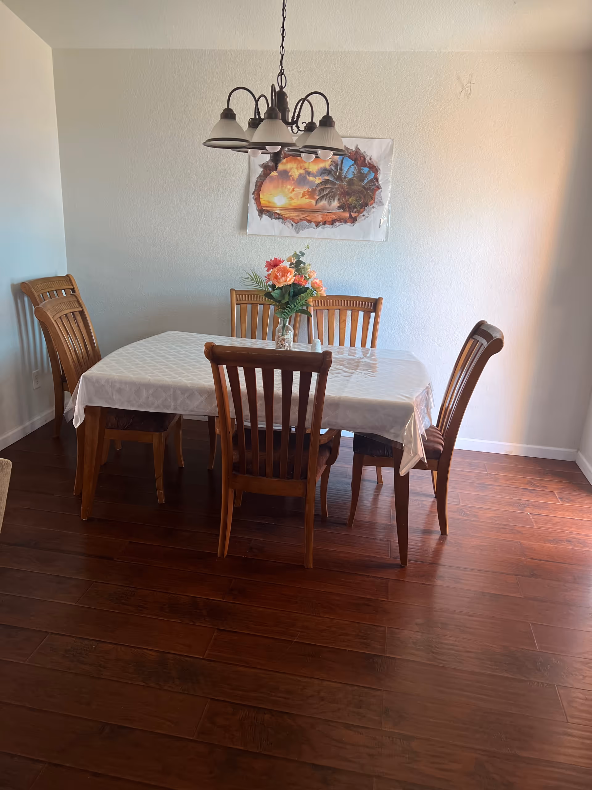 A dining room with a wooden table, chairs, chandelier, wall art, and a flower centerpiece.