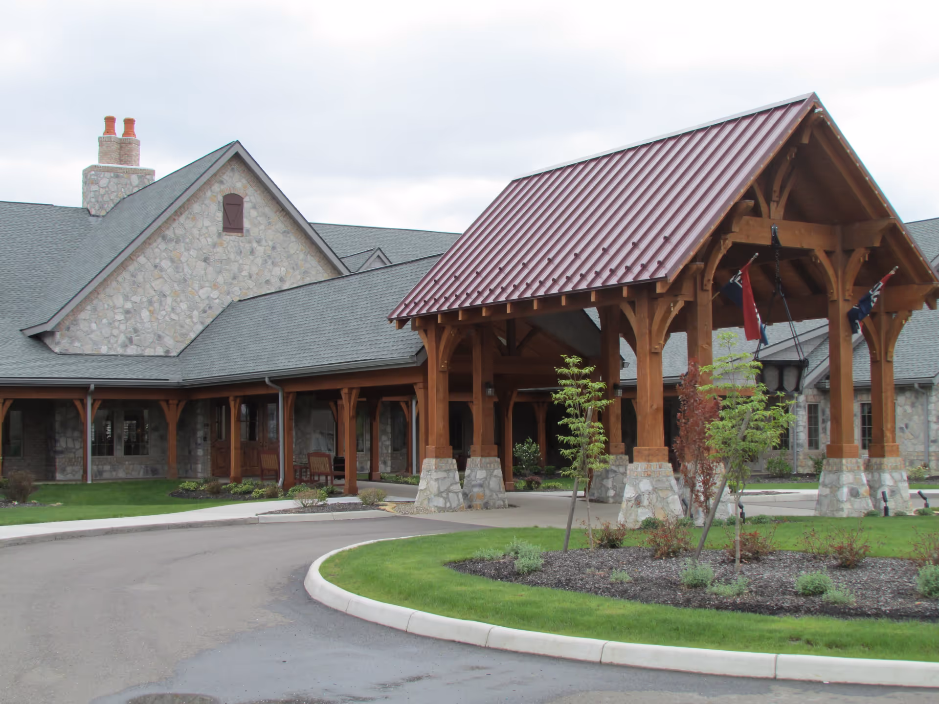 Front entrance of a stone-clad senior living building featuring a large timber porte-cochere with a red metal roof and a circular driveway.