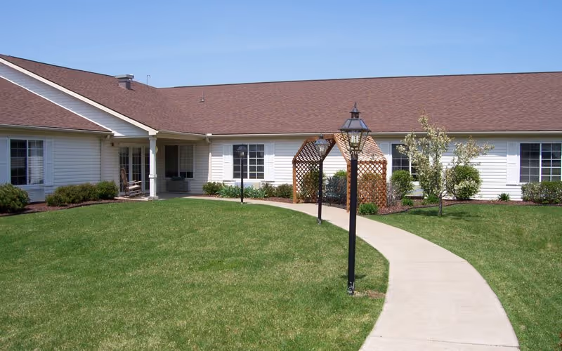 Single-story senior living facility exterior with curved walkway, lamppost, and well-kept lawn.