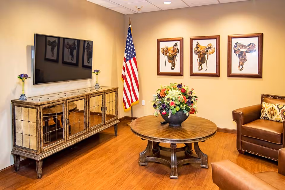 Welcoming lounge with a wall-mounted TV, antique console, round wooden table with a floral arrangement, leather chairs, and an American flag.