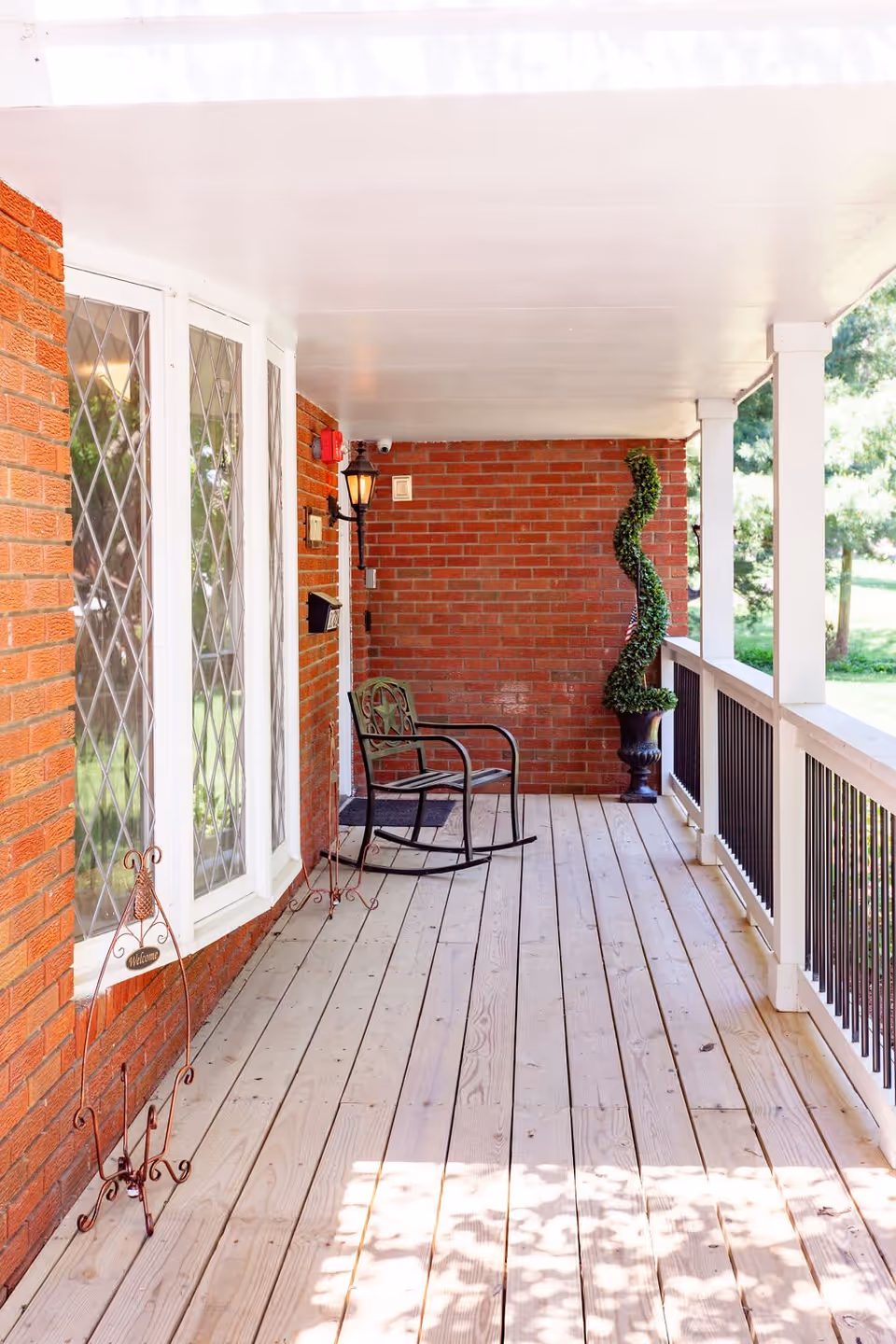 Covered front porch with wood plank flooring, a metal rocking chair, brick wall, bay windows and a spiral topiary in a planter.