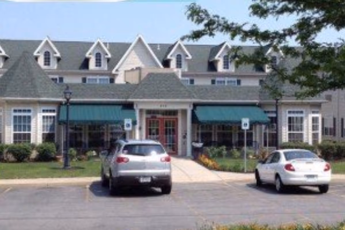 Exterior front view of Peregrine Senior Living at Cheektowaga building with a green awning over the entrance, two cars parked in front, and a clear blue sky.