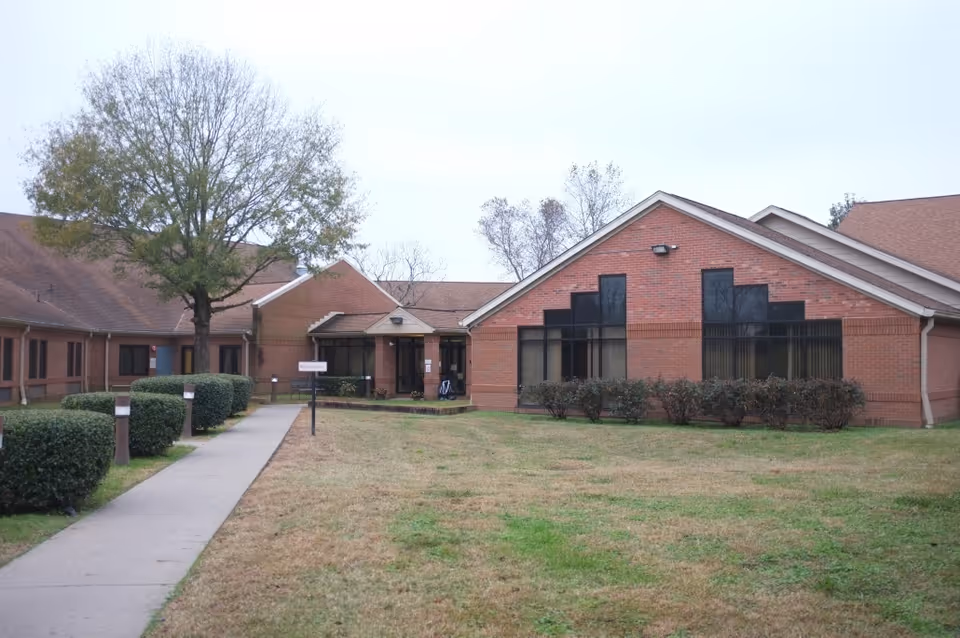 Exterior view of a single-story brick building with large windows and a walkway leading to the entrance. There are trimmed bushes along the walkway and a tree on the left side of the image. The sky is overcast.