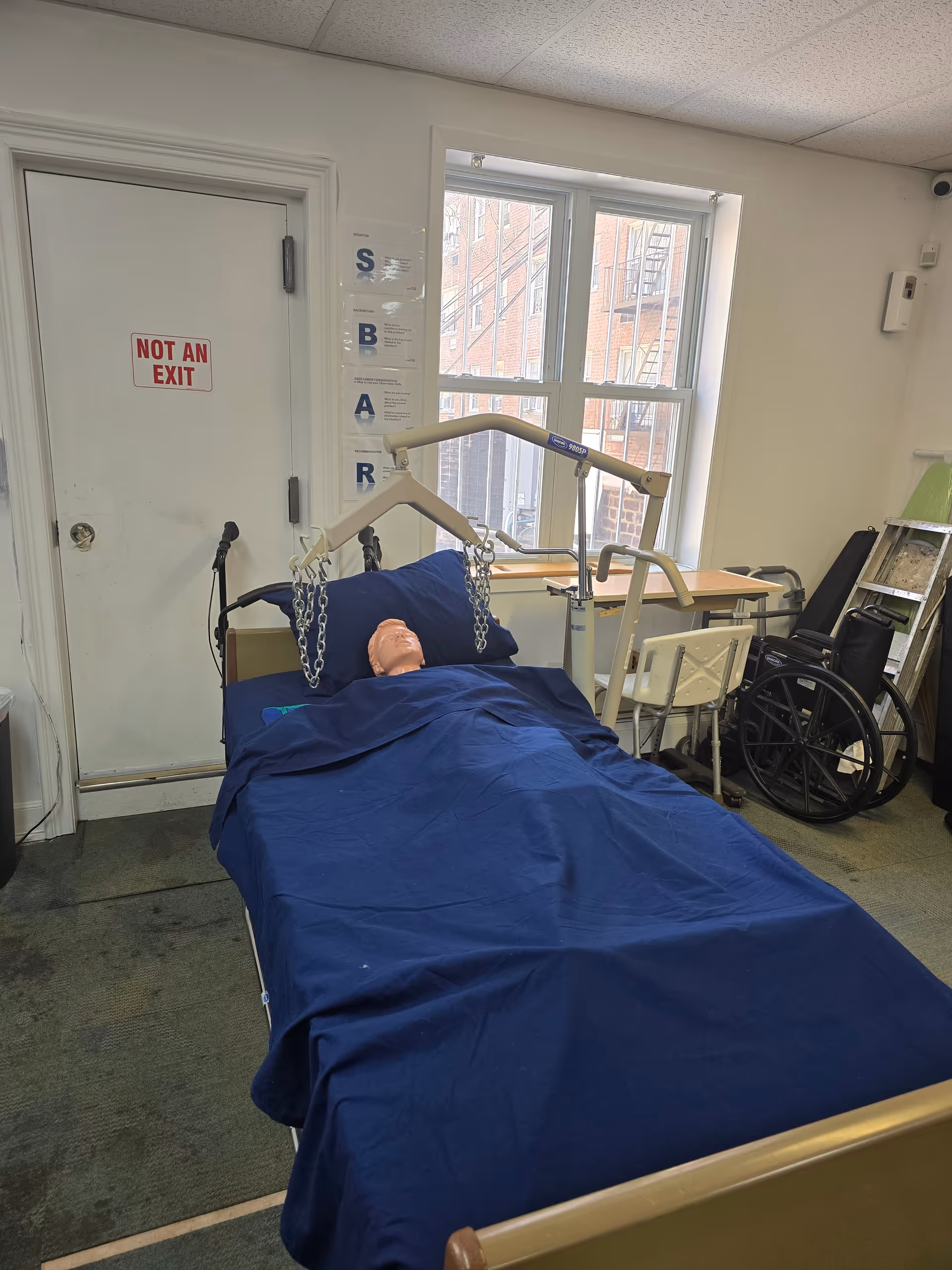 Hospital-style patient bed with blue sheets, trapeze bar, and wheelchair beside a window.