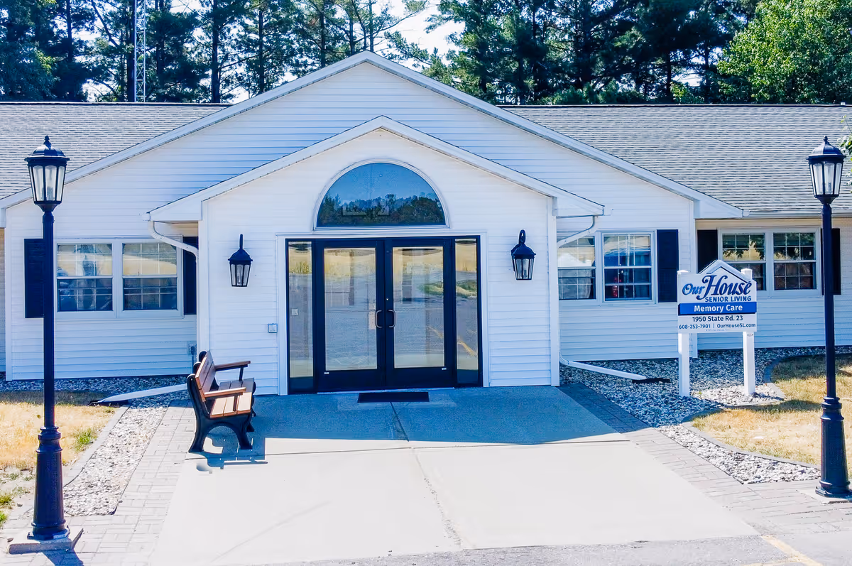 Front exterior view of a single-story white building with a peaked roof, black double doors with glass panels, two black wall-mounted lanterns on either side of the entrance, a bench on the left side of the walkway, two black lamp posts on either side of the walkway, and a sign on the right side that reads 'Our House Senior Living Memory Care, 1950 State Rd. 23, 608-253-7901, OurHouseSL.com'. Trees are visible in the background.