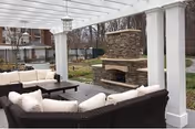 Outdoor lounge area with white cushioned wicker seating and a stone fireplace beneath a pergola.