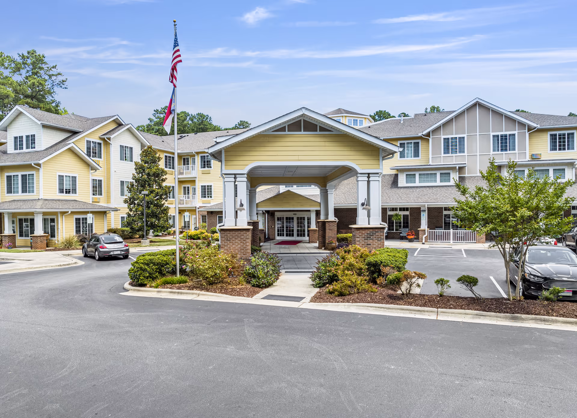 Front exterior view of Whispering Pines Gracious Retirement Living facility showing a large covered entrance with columns, landscaped bushes, an American flag on a flagpole, parked cars, and a multi-story building with yellow and beige siding under a blue sky.