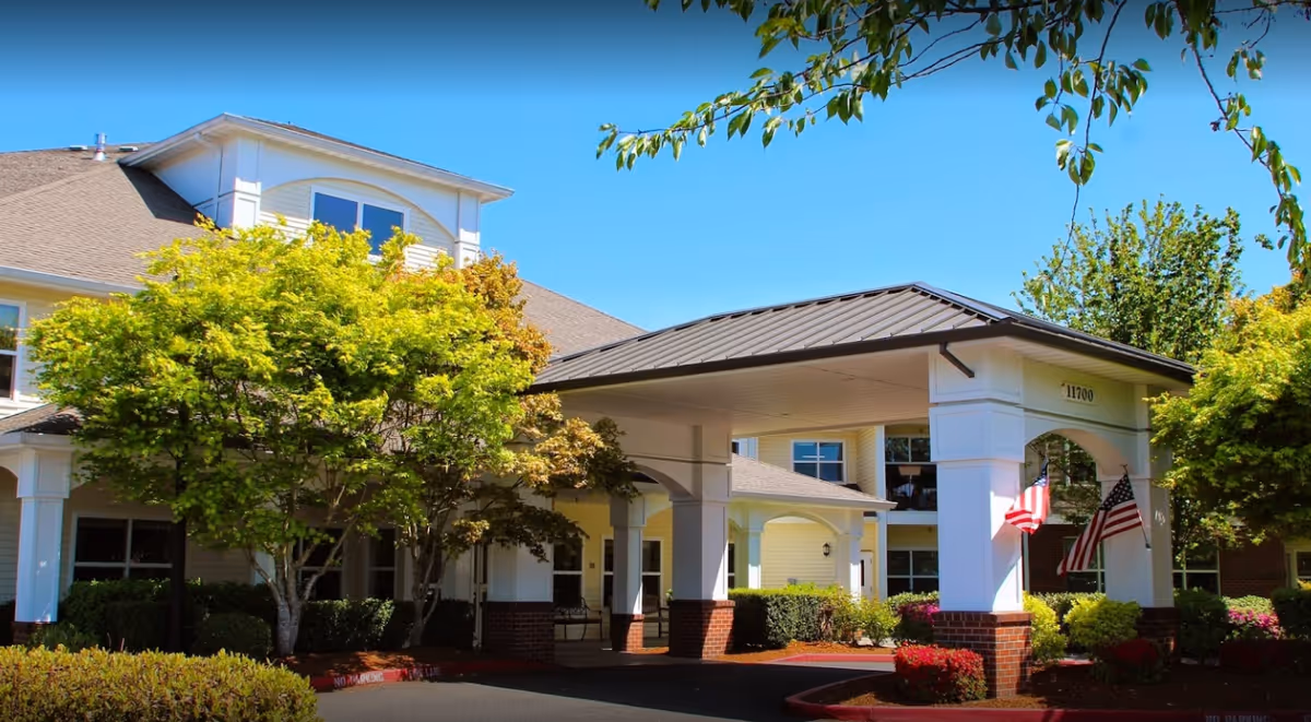 Exterior view of Prestige Senior Living Bridgewood showing the entrance with a covered driveway supported by white columns with brick bases. The building has light yellow siding and a gray roof. There are green trees and well-maintained landscaping with bushes and flowers around the entrance. Two American flags are displayed on the right side of the entrance.