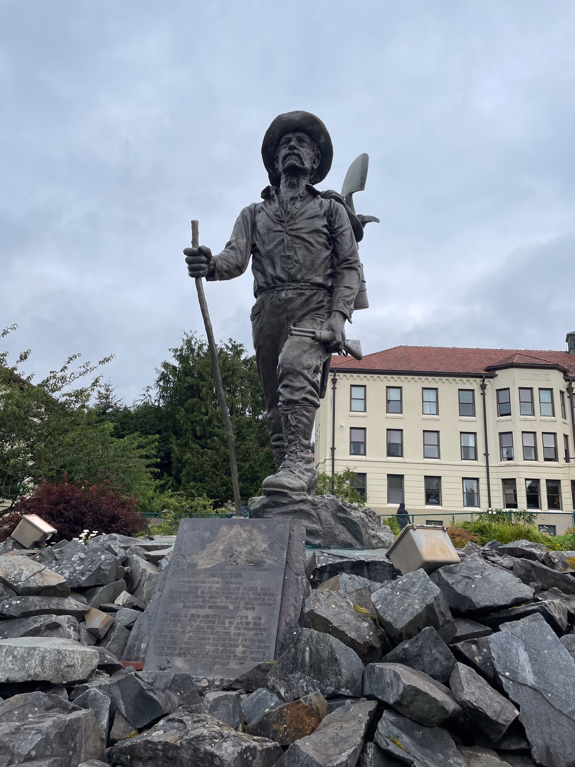 A statue of a pioneer with a shovel stands on rocks in front of beige building.