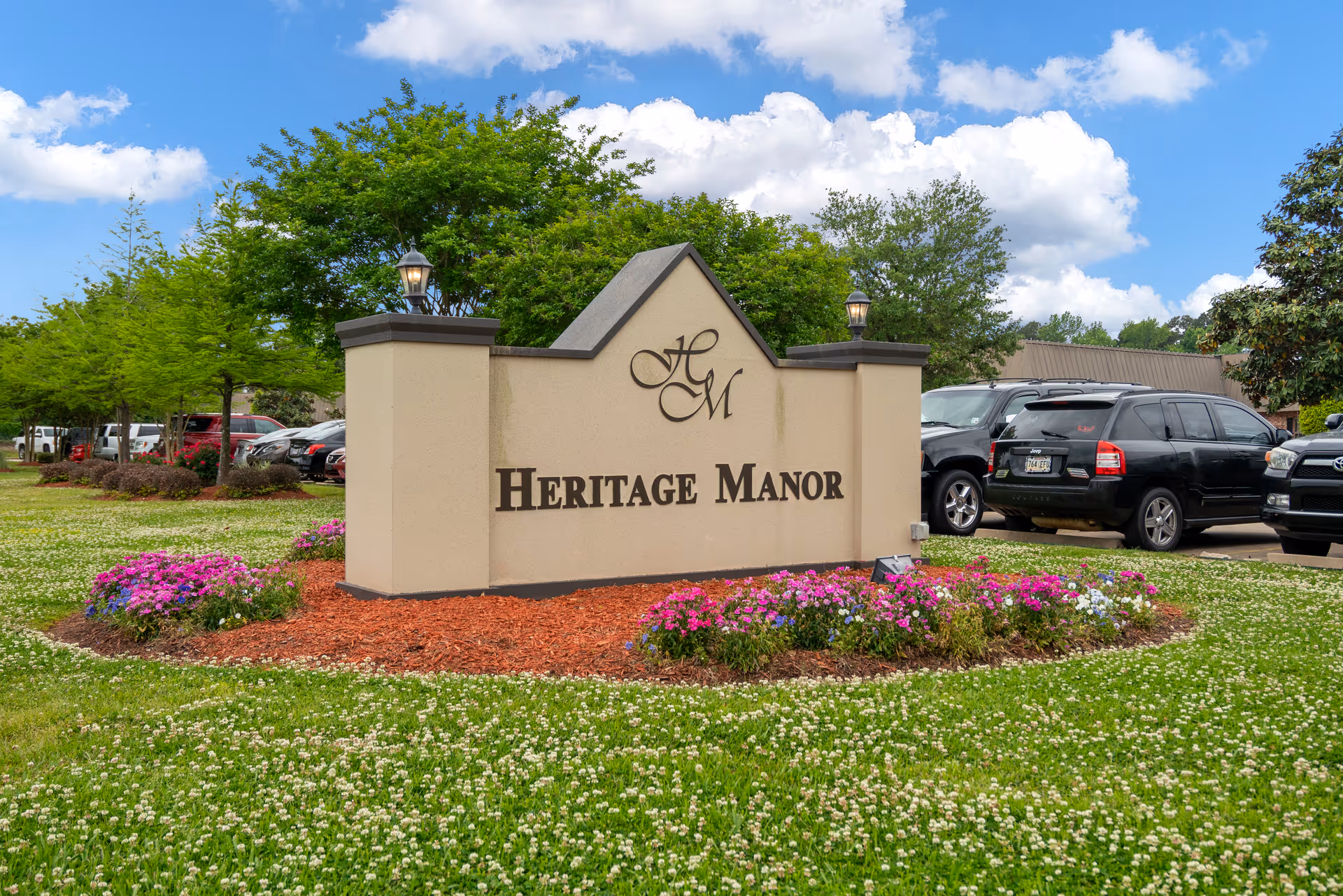 A large beige sign with the letters HM and the words Heritage Manor in black, surrounded by a landscaped area with flowers and mulch. Behind the sign, there are several parked cars and green trees under a partly cloudy blue sky.