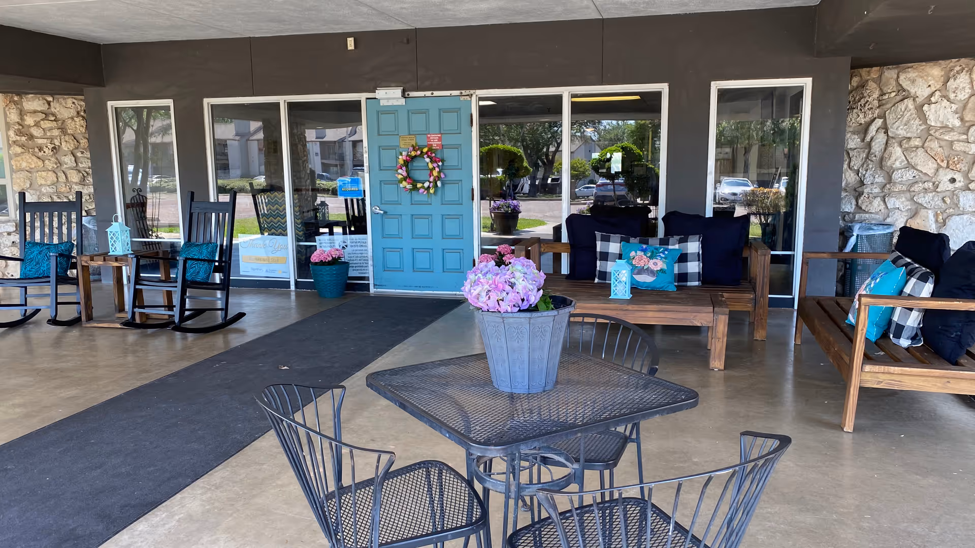 Covered entrance patio of a healthcare center with a blue door, seating area of benches and rocking chairs, metal table and chairs, and potted flowers.