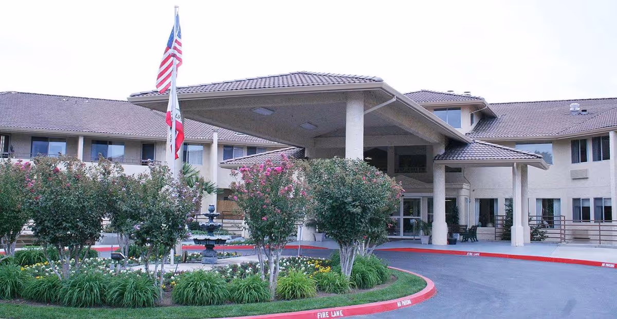 Front entrance of a senior living facility with a covered porte-cochère, flagpoles, landscaped circular driveway and a fountain.