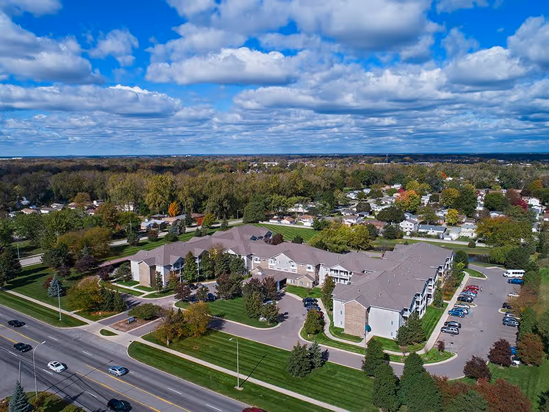 Aerial view of a large senior living facility named American House Southland Senior Living surrounded by trees and residential houses, with a parking lot and cars visible on the right side, under a partly cloudy sky.