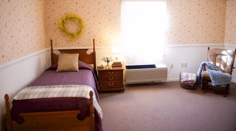 A cozy bedroom with a single wooden bed covered in a purple quilt, a nightstand with a lamp beneath a window and an armchair in the corner.