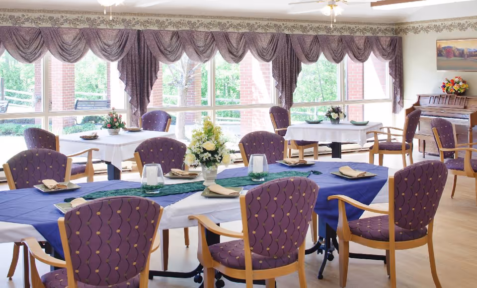 Bright dining room with several tables set with purple upholstered chairs, floral centerpieces, and large windows overlooking greenery.
