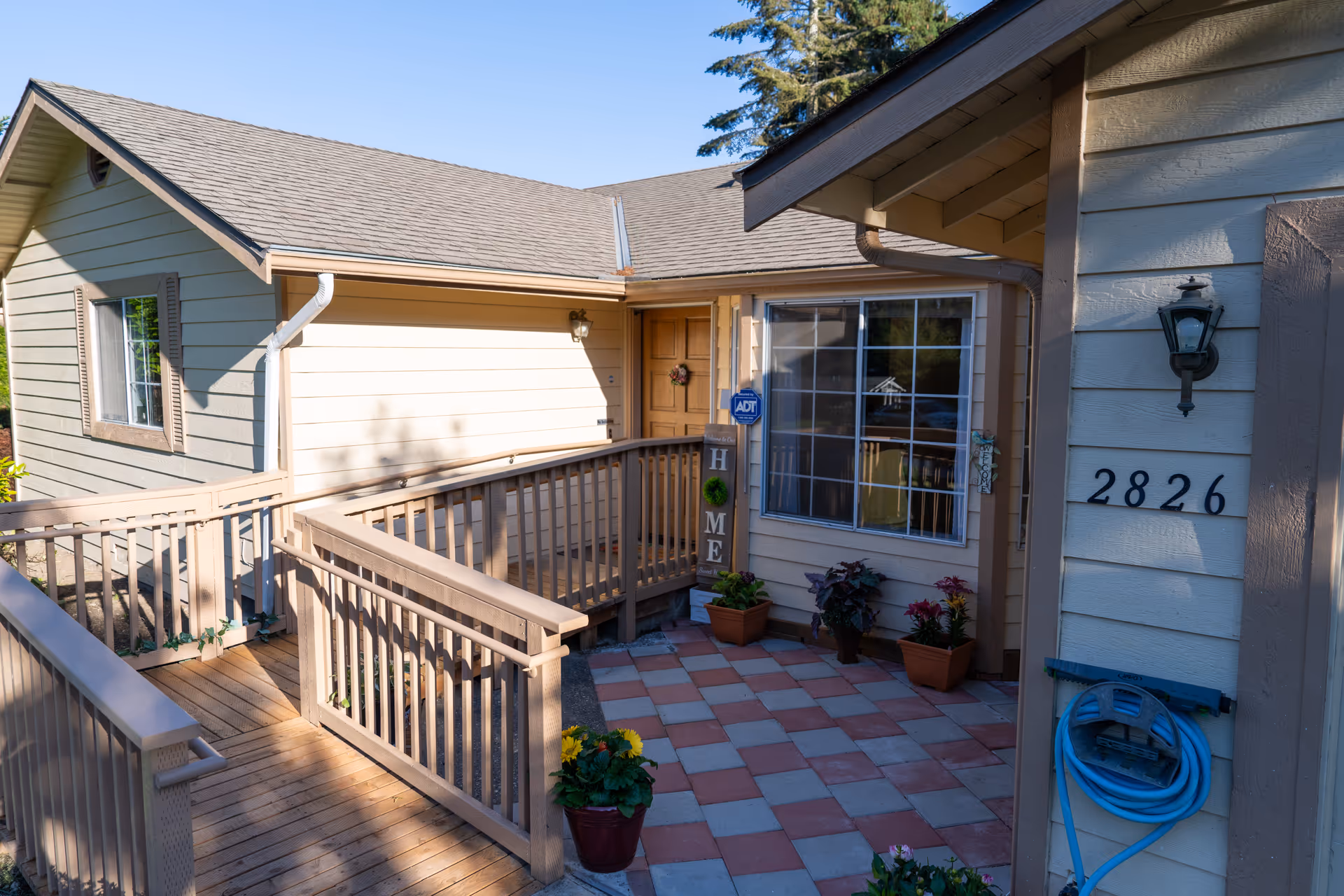 Front exterior of a senior living home with a wooden railing, tiled porch, and plants.