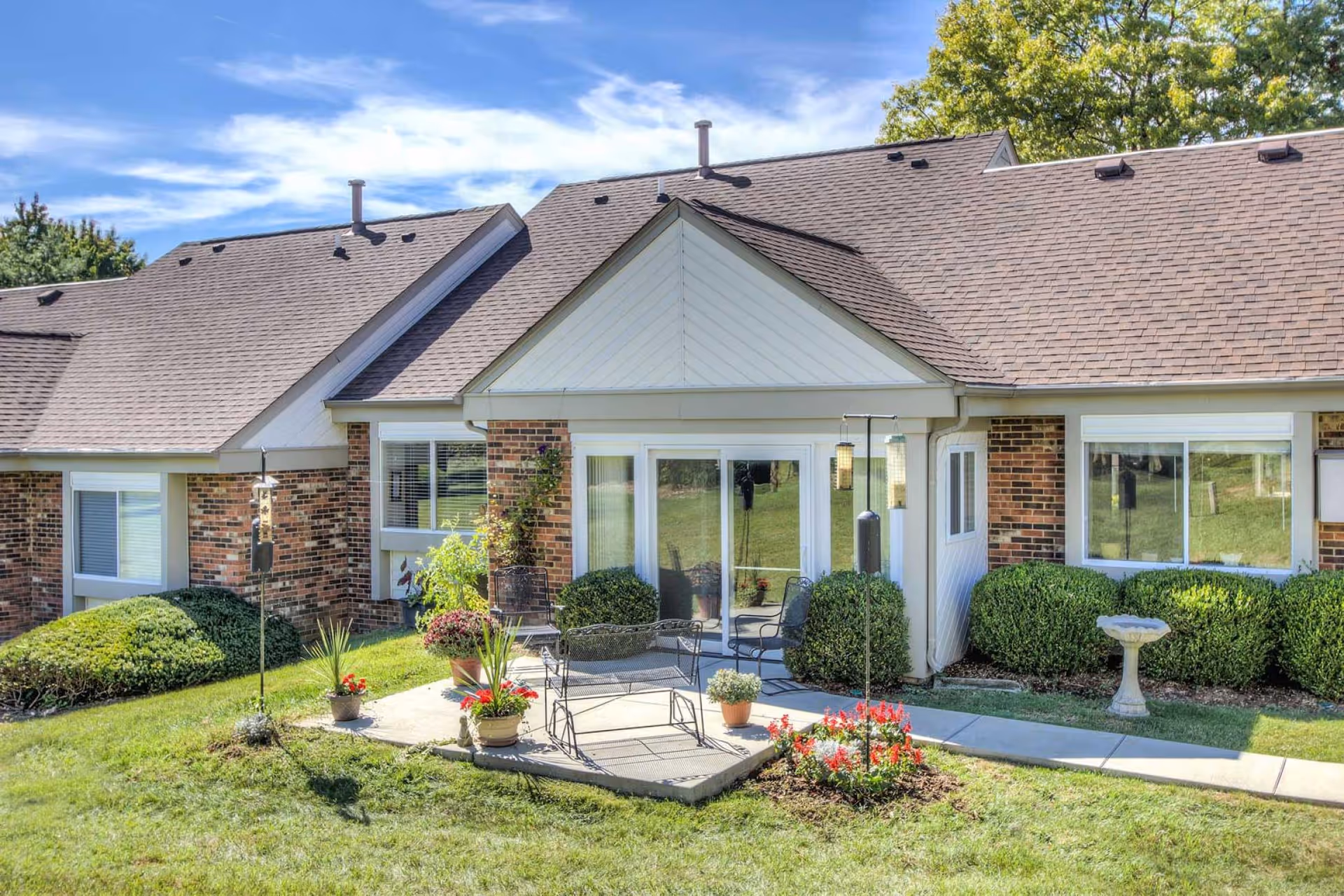Front exterior of a brick senior living unit with a small patio, chairs, potted plants, and a manicured lawn.