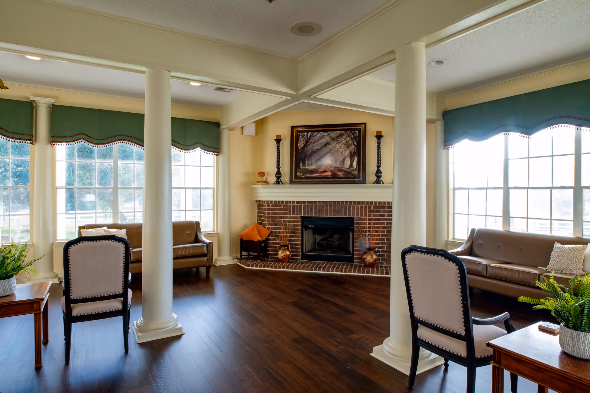 A bright and spacious living room with large windows covered by green valances, two brown leather sofas, two upholstered chairs, a wooden side table with a potted plant, and a brick fireplace with decorative items and a framed picture above it.