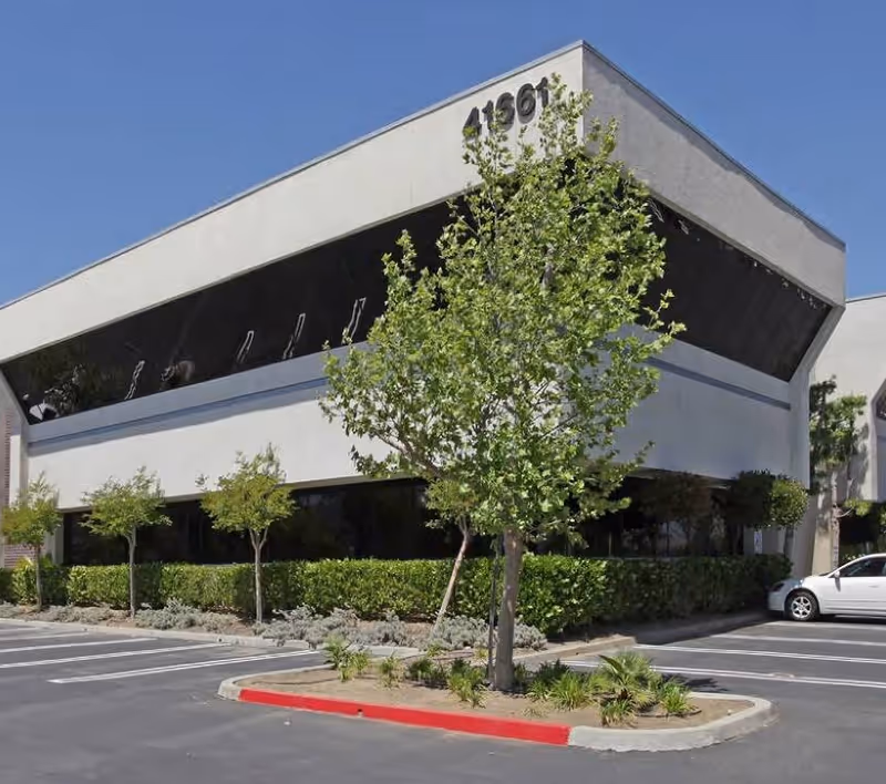 White modern building with an angled upper facade, trimmed trees, and a parking lot.