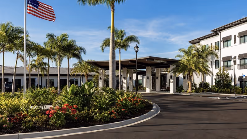 Exterior view of American House Naples senior living facility with palm trees and flag.